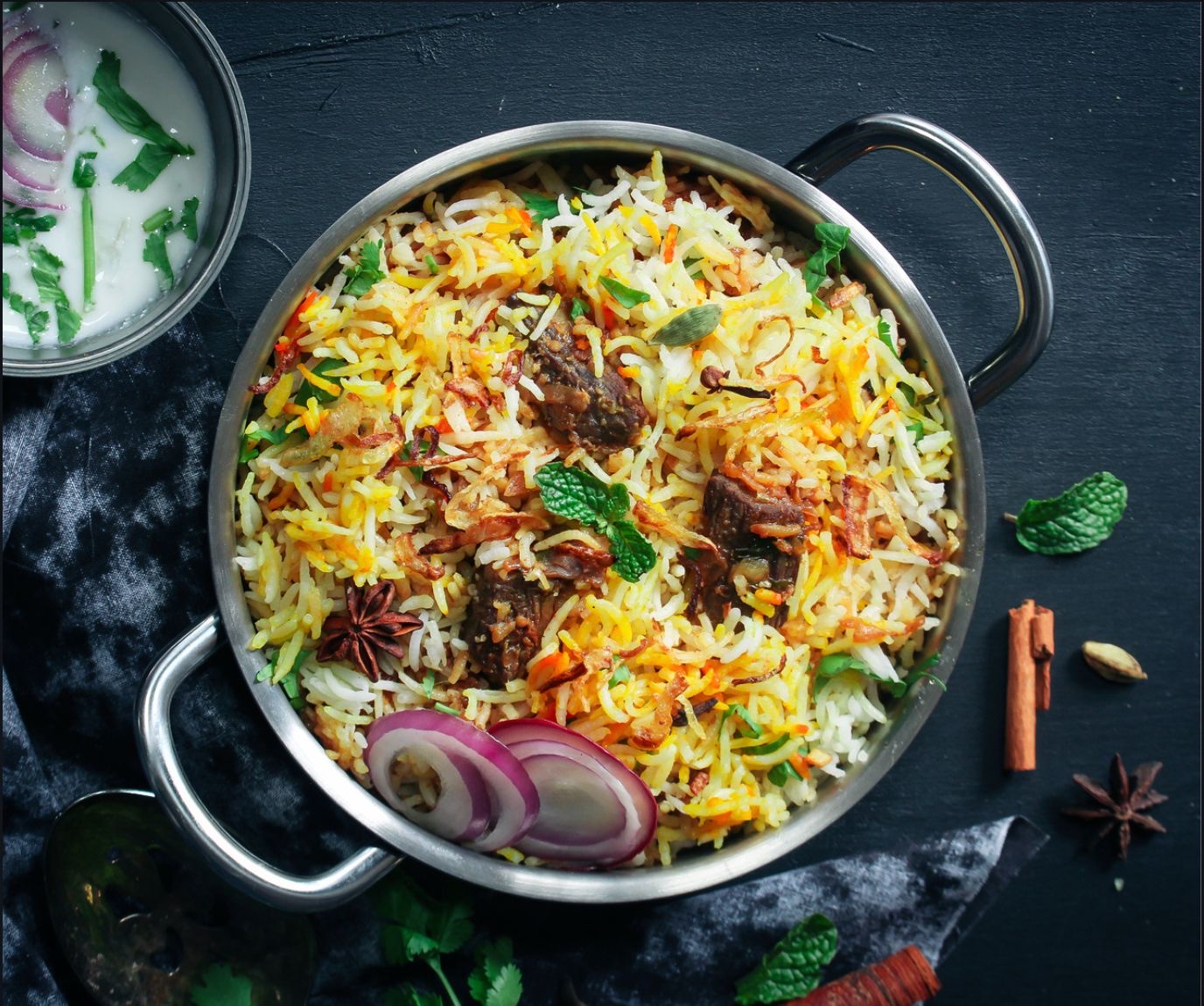 Hyderabadi dum biryani in a steel handi with raita, whole spices and onion